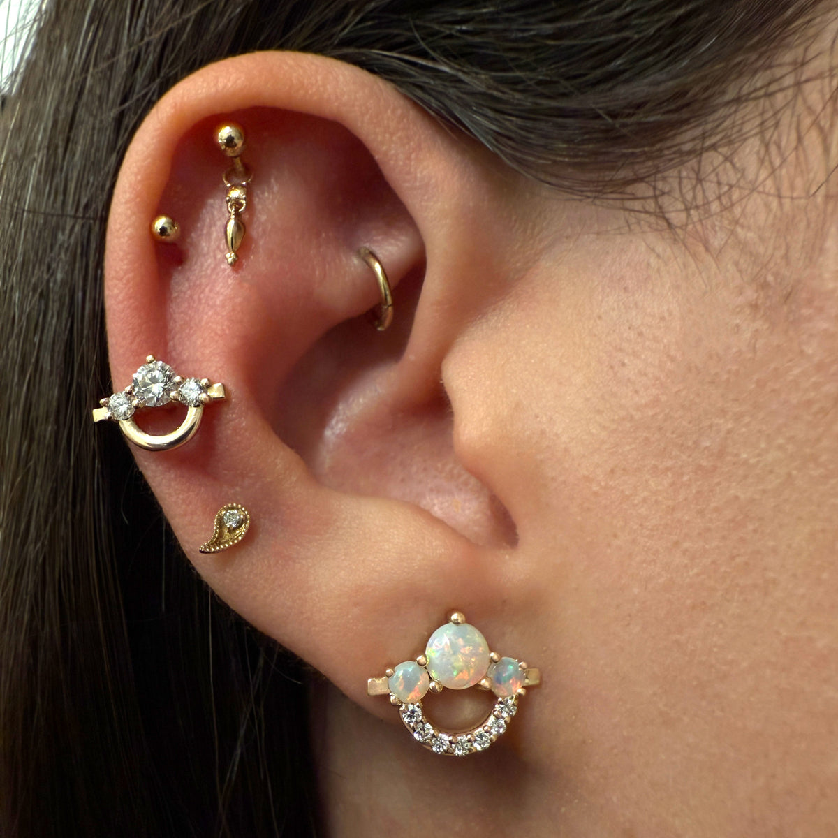 Close-up of a person's ear showcasing multiple gold-toned ear piercings, including a hoop with a clear stone, several small studs, a curved hoop, and a crescent moon stud with an opal accent.