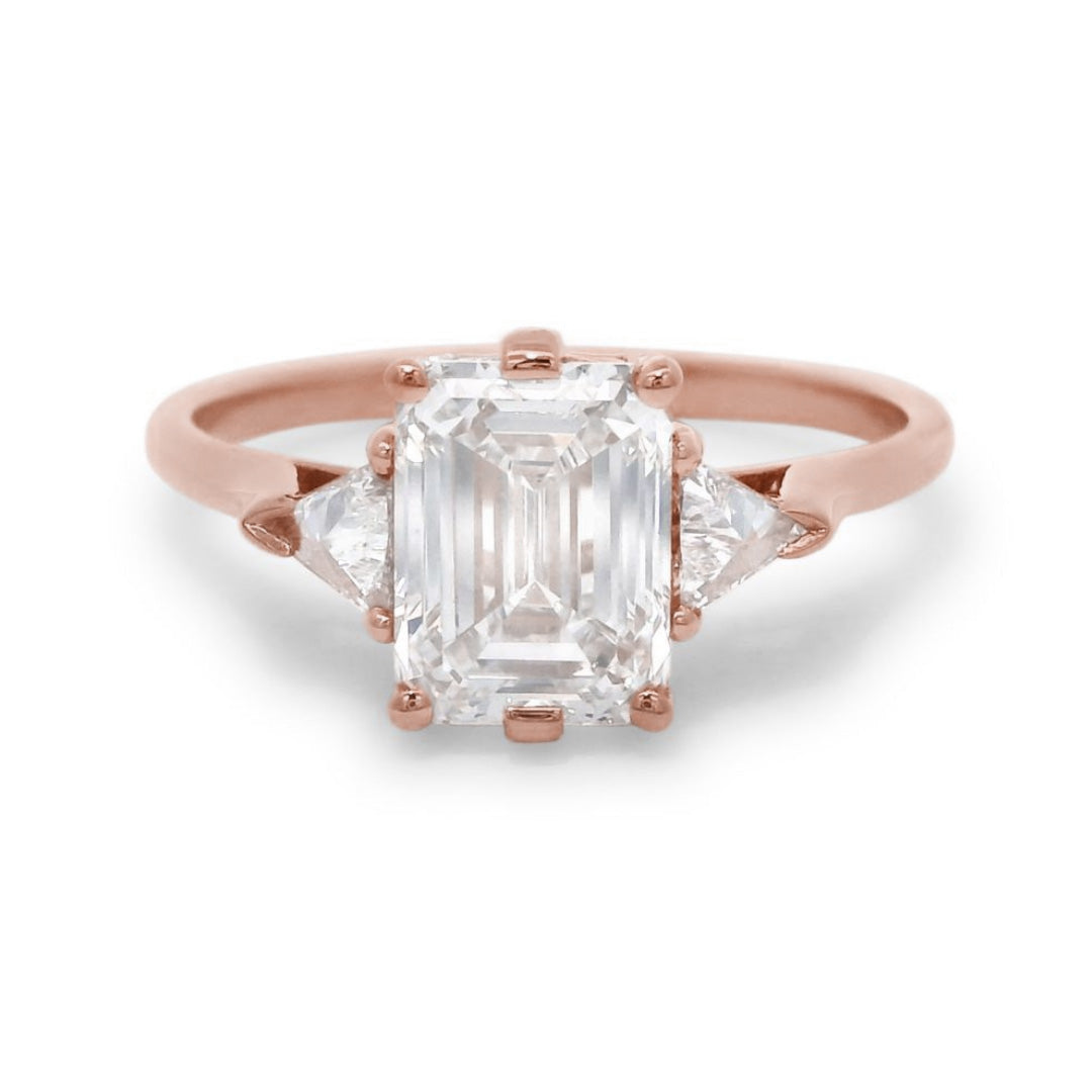 A rose gold three stone engagement ring featuring an emerald cut diamond center and two trillion diamond side stones on a white background
