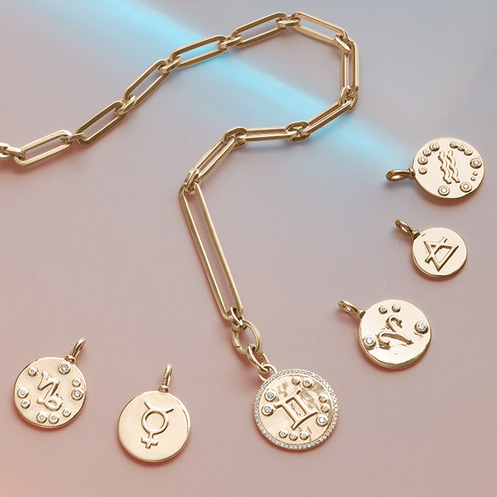 A cascade of yellow gold zodiac charms next to a mixed link chain on a pink background.
