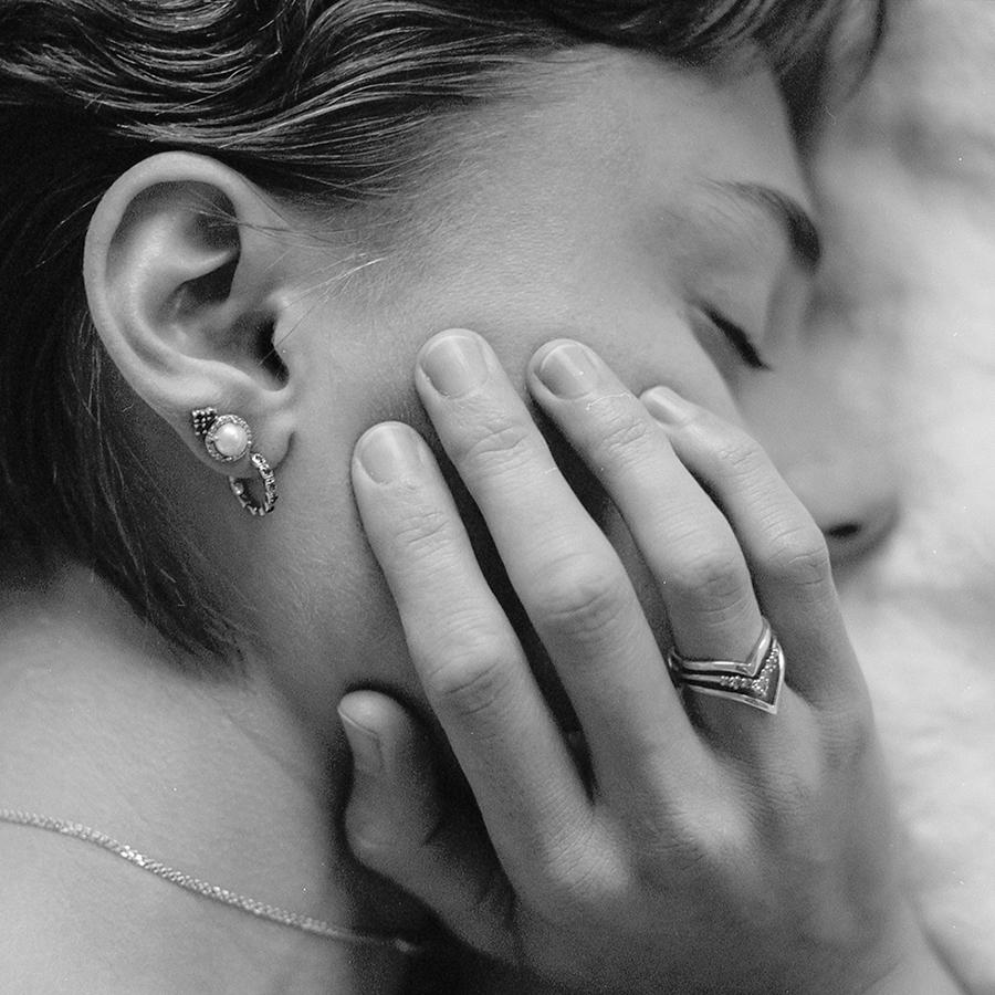 Black and white photo of a model laying down wearing two stud earrings and one huggie hoop and touching her face with her right hand. On her right ring finger she wears three nesting bands.