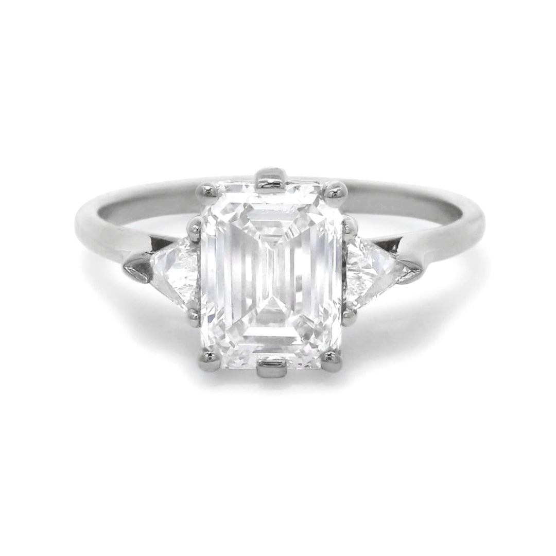 A white gold three stone engagement ring featuring an emerald cut diamond center and two trillion diamond side stones on a white background