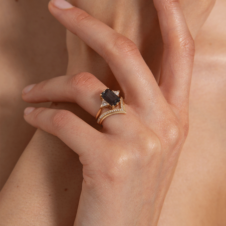 A three stone engagement ring with an emerald shape grey spinel center stone with two white diamond trillion side stones set in yellow gold above a chrysalis band with white diamonds set in yellow gold on a medium-complexion model. Available in White Gold, Yellow Gold, Rose Gold.