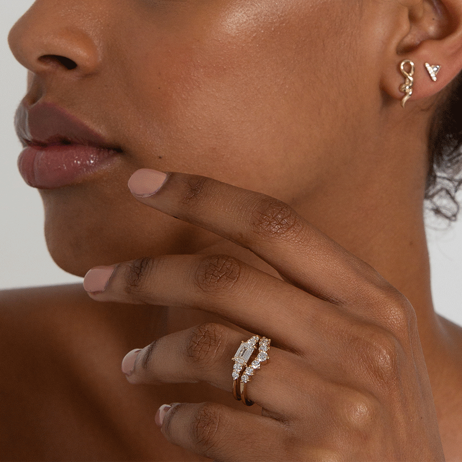 A ring with an emerald cut and shaped white diamond center stone with two round white diamonds on either side set in yellow gold set above an 11 white diamond curve band on a darker-complexion model.