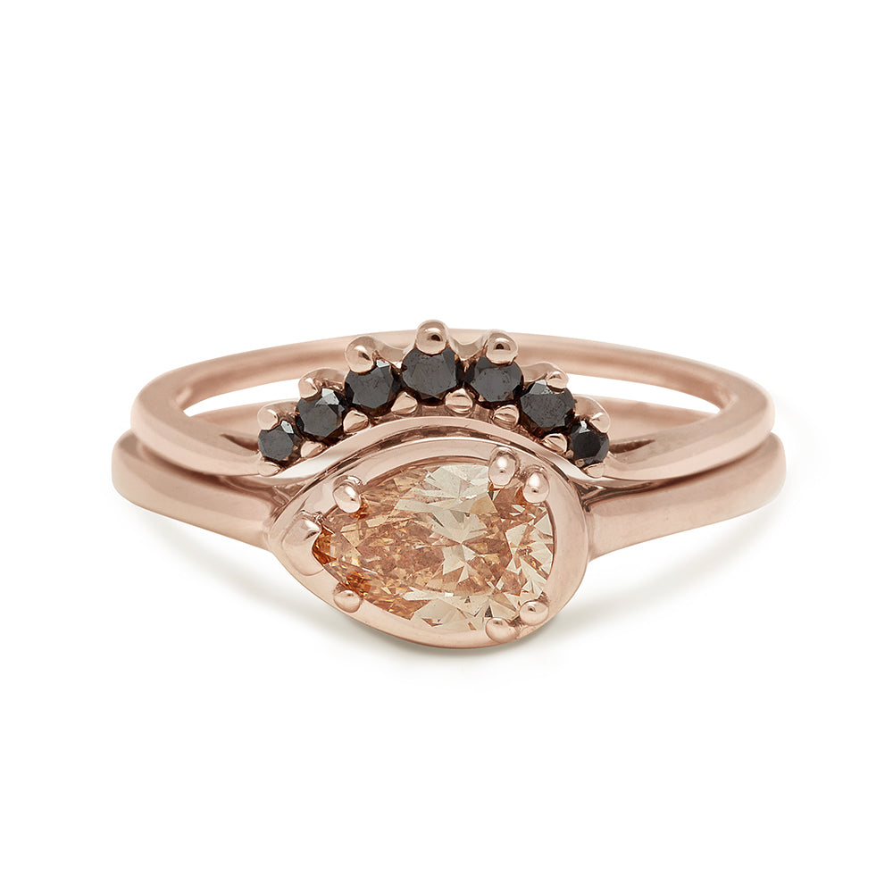 A pear shaped ring with a champagne diamond set in rose gold stacked above a ​​curved band with 7 black diamonds in ascending and descending size set in rose.