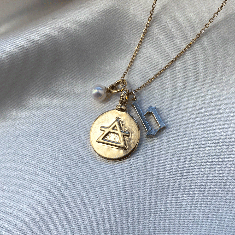 A 14mm charm with the air element sign set in yellow gold on a yellow gold chain with an initial charm and a pearl charm.