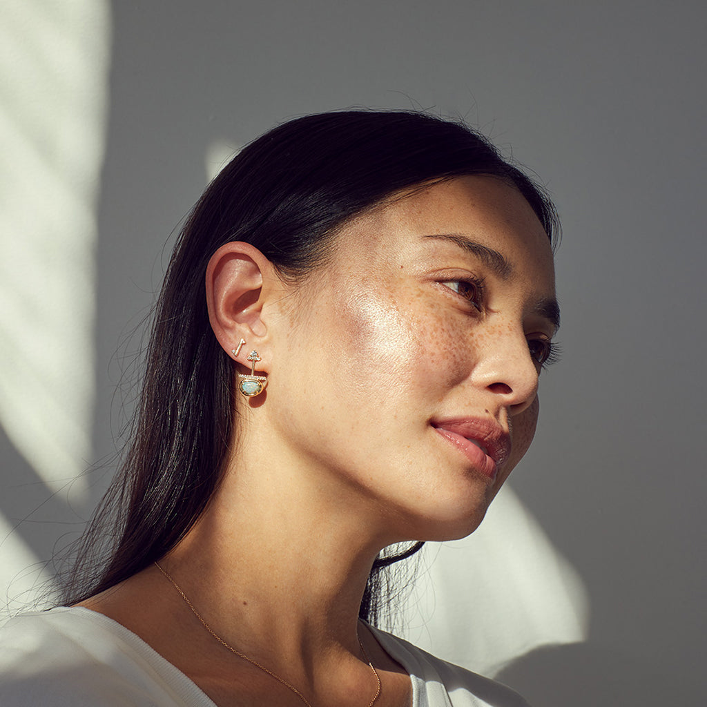 A single straight line earring with white diamond accent stones on either side set in yellow gold next to an amulet earring on a medium-complexion model.