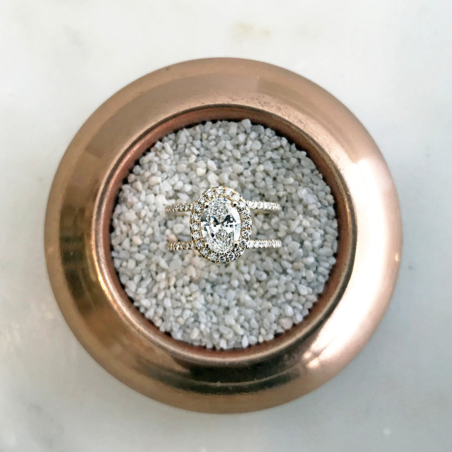 A double banded bezel set oval white diamond ring with a white diamond halo and white diamond pave around the band set in yellow gold in a bowl with white stones.