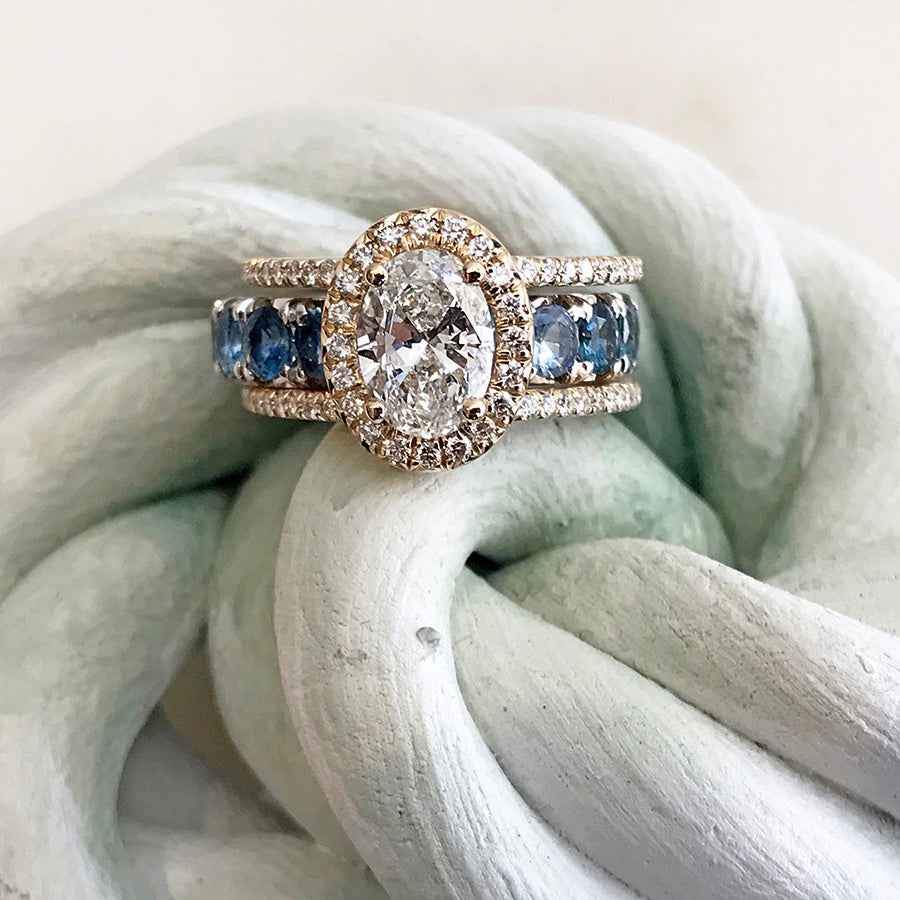 A double banded bezel set oval white diamond ring with a white diamond halo and white diamond pave around the band set in yellow gold with a blue gemstone eternity band set in white gold on a white fabric background.