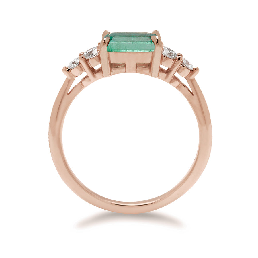 Side view of a ring with an emerald cut and shaped green emerald center stone with two round white diamonds on either side set in rose gold., Rose Gold
