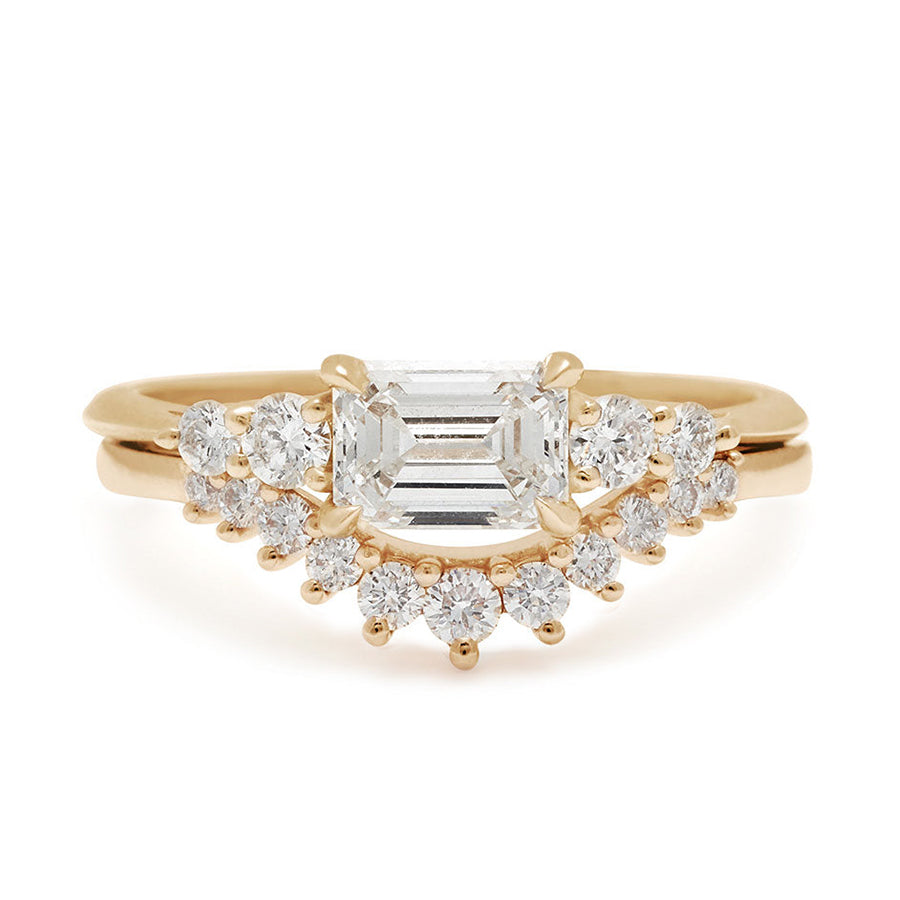 A ring with an emerald cut and shaped white diamond center stone with two round white diamonds on either side set in yellow gold set above an 11 white diamond curve band.