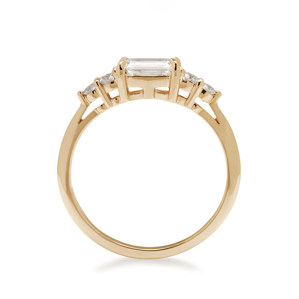 A side view of a ring with an emerald cut and shaped white diamond center stone with two round white diamonds on either side set in yellow gold., Yellow Gold