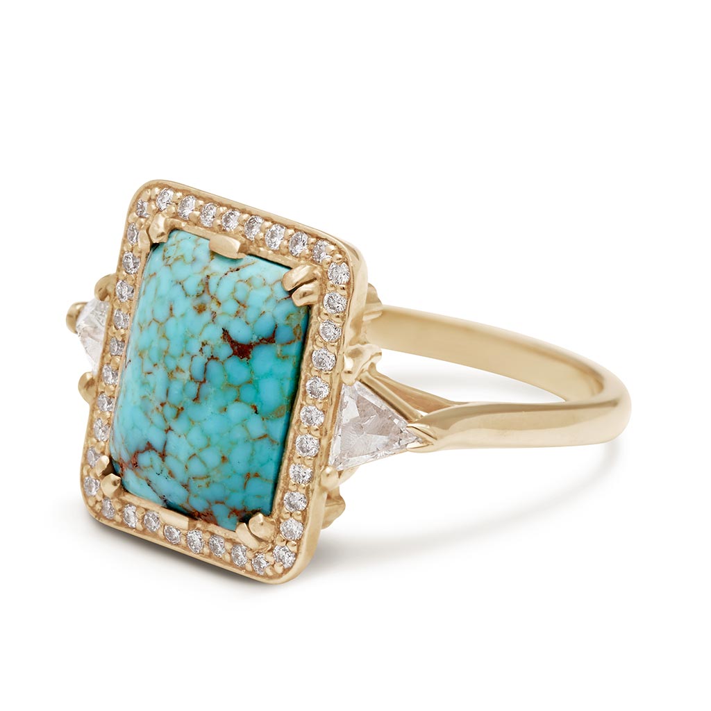Angled view of a one of a kind three stone engagement ring with an emerald shape turquoise center stone surrounded by a white diamond pave halo with two white diamond trillion side stones set in yellow gold.