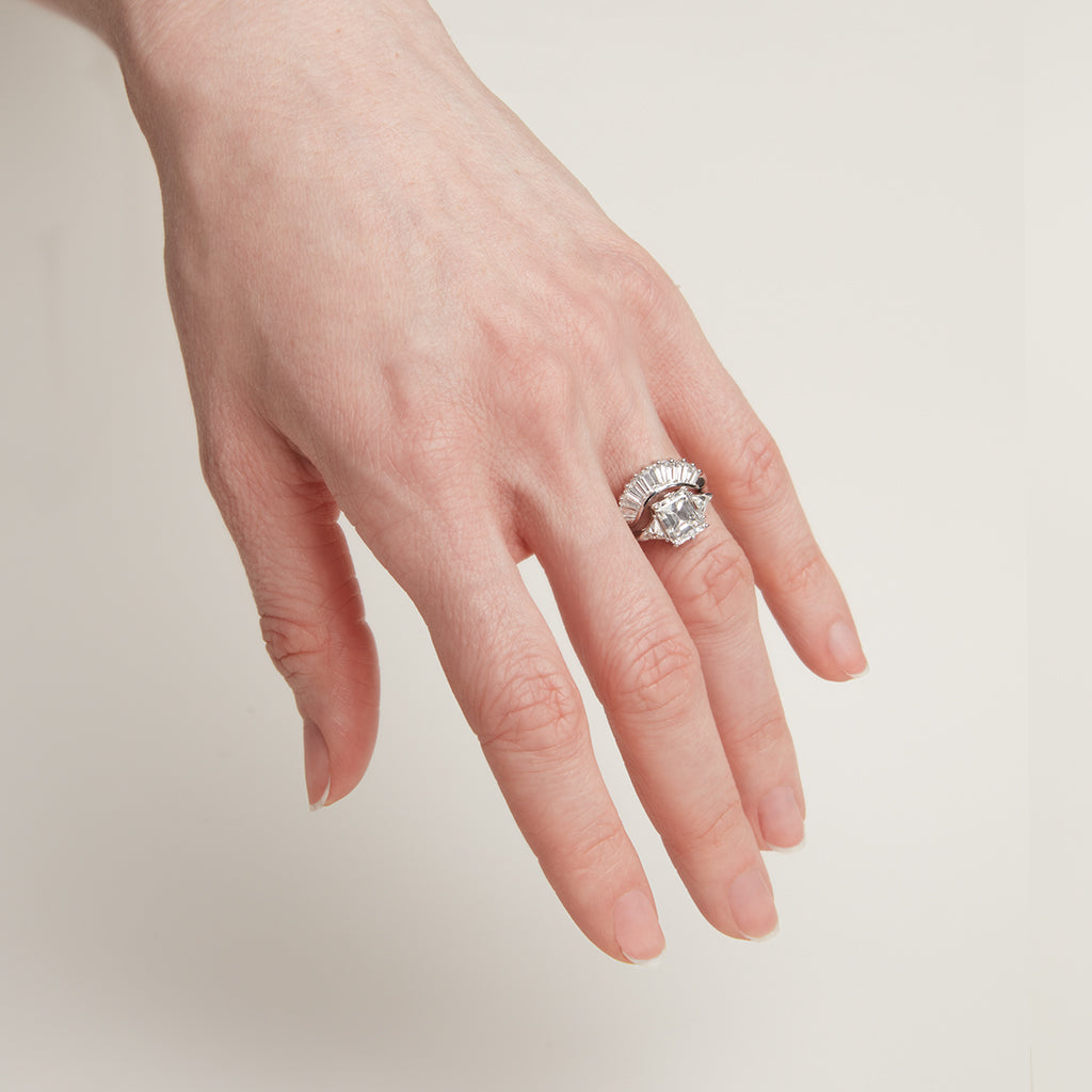 11 white diamond baguettes set in a curved band and in white gold stacked above a three stone engagement ring with an emerald cut and emerald shape white diamond center stone with two white diamond trillion side stones set in white gold on a light-complextion hand on a pink background.