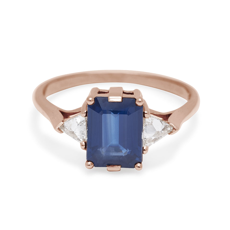 A three stone engagement ring with an emerald cut and emerald shape 8x6mm blue sapphire center stone with two white diamond trillion side stones set in rose gold., Rose Gold