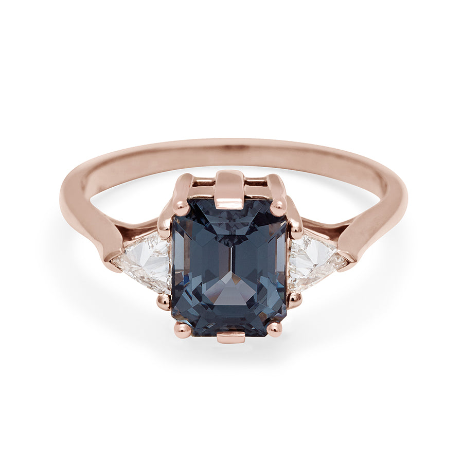 A three stone engagement ring with an emerald cut and emerald shape grey spinel center stone with two white diamond trillion side stones set in rose gold., Rose Gold