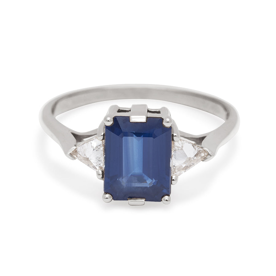 A three stone engagement ring with an emerald cut and emerald shape 8x6mm blue sapphire center stone with two white diamond trillion side stones set in white gold., White Gold
