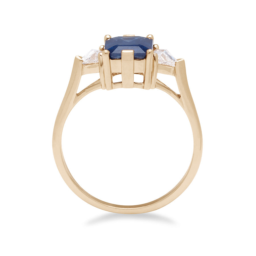 Side view of a three stone engagement ring with an emerald cut and emerald shape 8x6mm blue sapphire center stone with two white diamond trillion side stones set in yellow gold., Yellow Gold