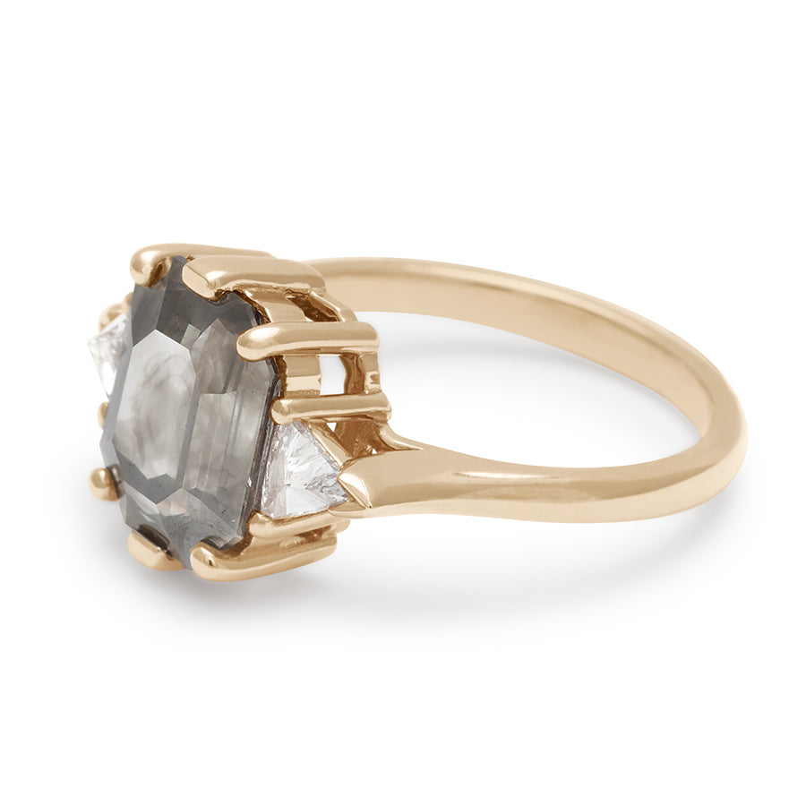 Angled view of a three stone engagement ring with an emerald cut and emerald shape grey diamond center stone with two white diamond trillion side stones set in yellow gold.