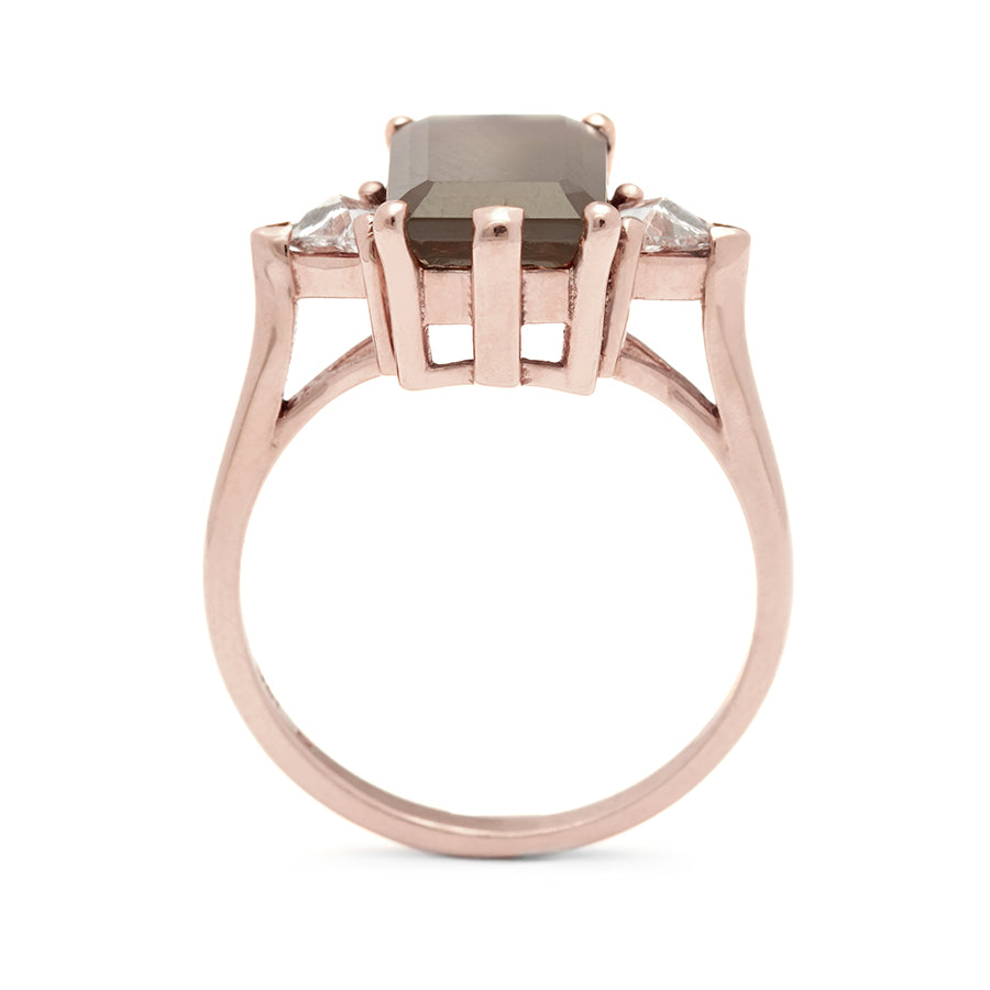 A side view of a three stone engagement ring with an emerald shape grey moonstone center stone with two white diamond trillion side stones set in rose gold., Rose Gold