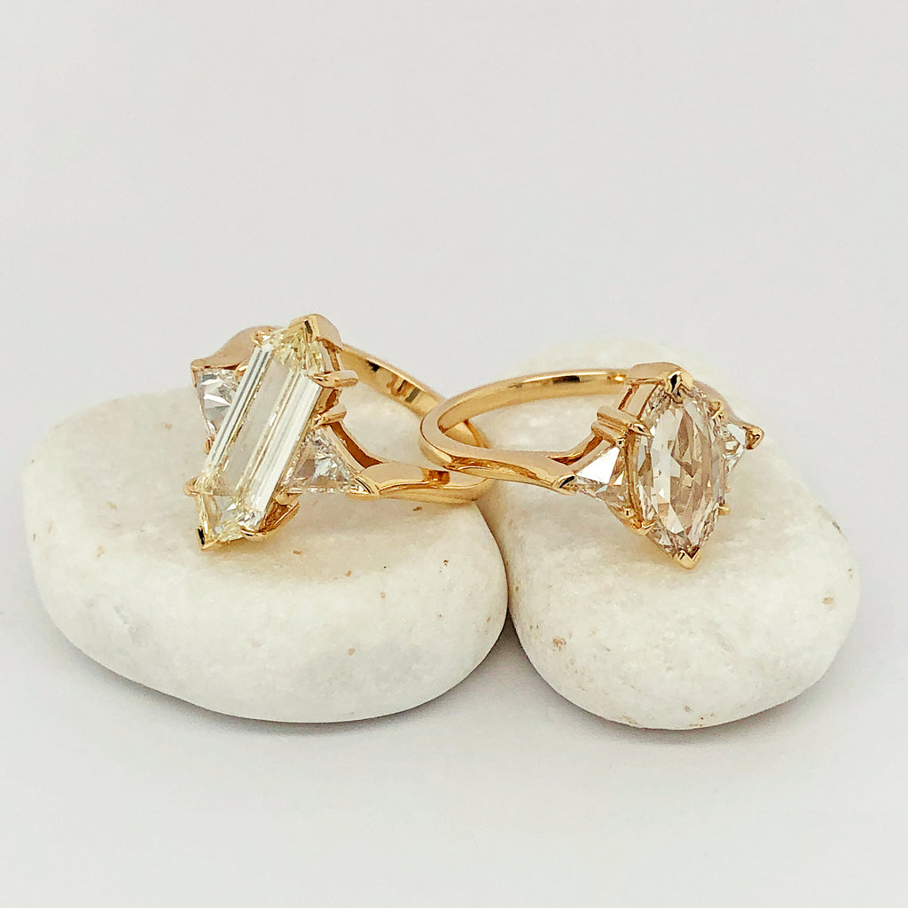 A three stone engagement ring with an hexagonal shape white diamond center stone with two white diamond trillion side stones set in yellow gold and a marquise shaped three stone ring on a white rock.