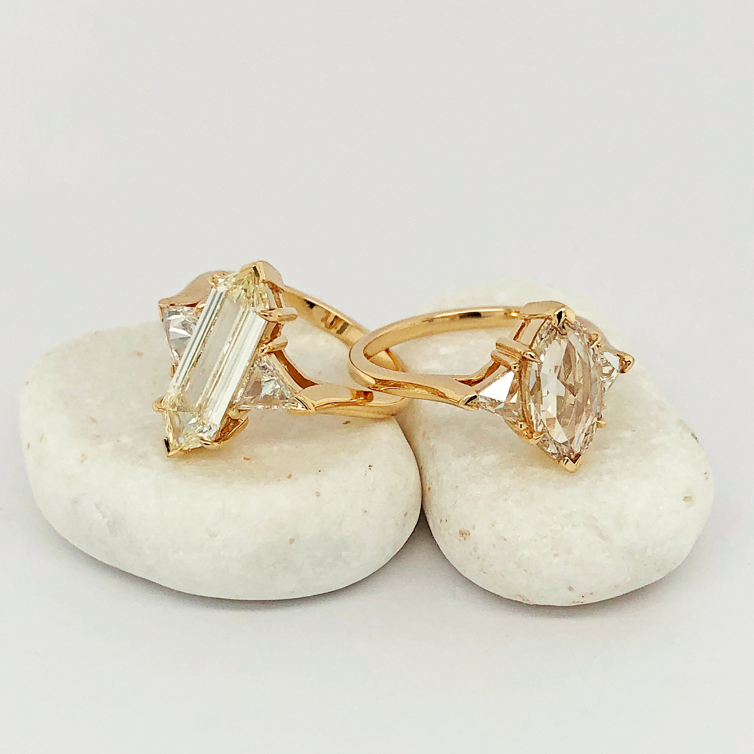 A three stone engagement ring with an hexagonal shape white diamond center stone with two white diamond trillion side stones set in yellow gold and a marquise shaped three stone ring on a white rock.