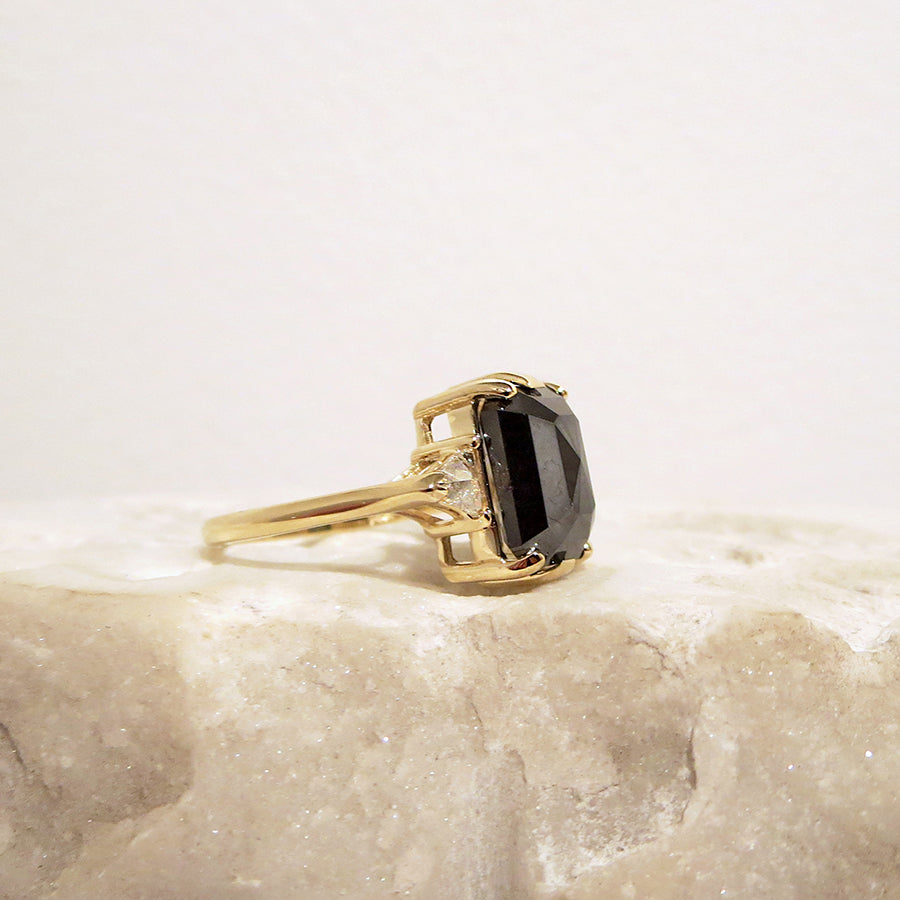 Side view of a three stone engagement ring with an emerald shape black diamond center stone with two white diamond trillion side stones set in yellow gold on a white rock.
