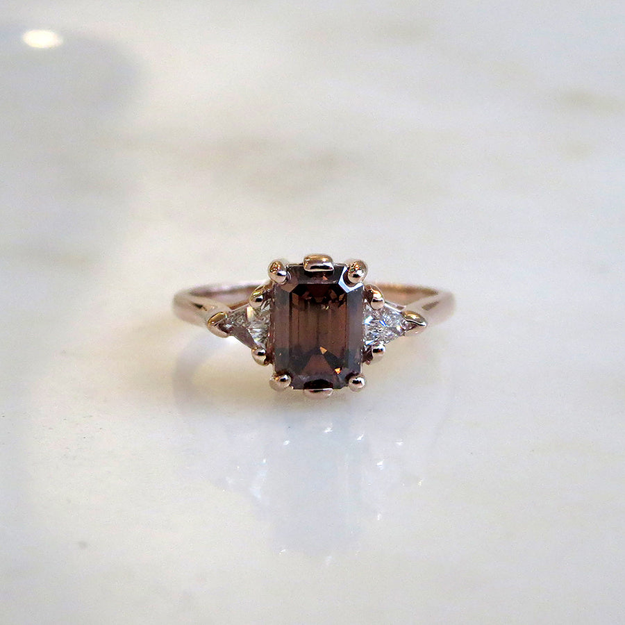 A three stone engagement ring with an emerald shape champagne diamond center stone with two white diamond trillion side stones set in yellow gold on a grey background.