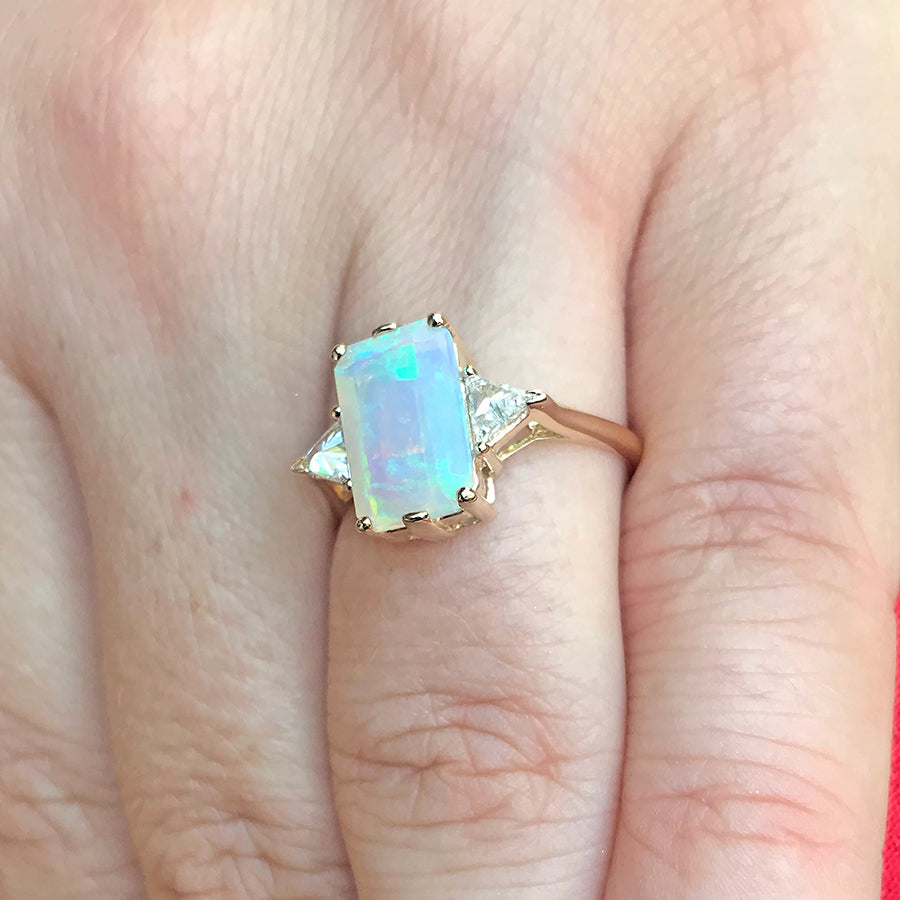 A three stone engagement ring with an emerald shape opal center stone with two white diamond trillion side stones set in yellow gold on a light-complextion hand.