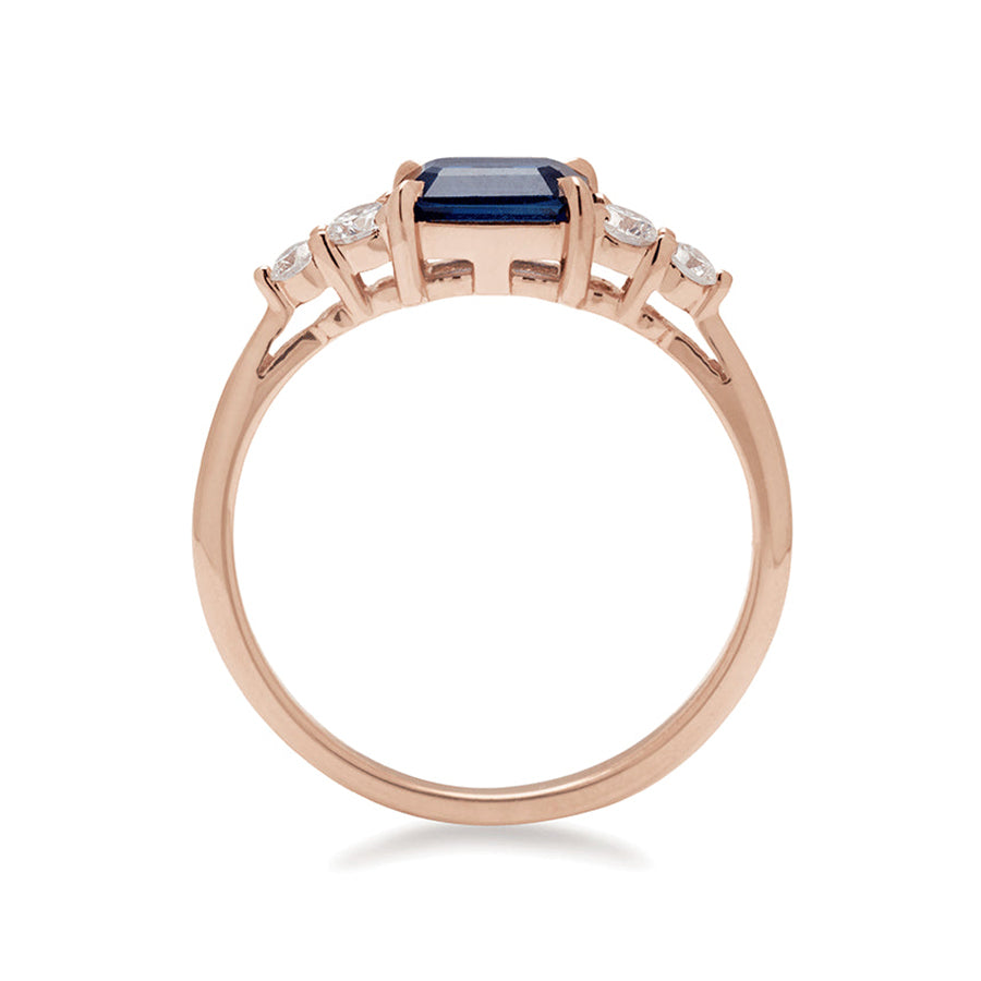 Side view of a ring with an emerald cut and shaped blue sapphire center stone with two round white diamonds on either side set in rose gold., Rose Gold