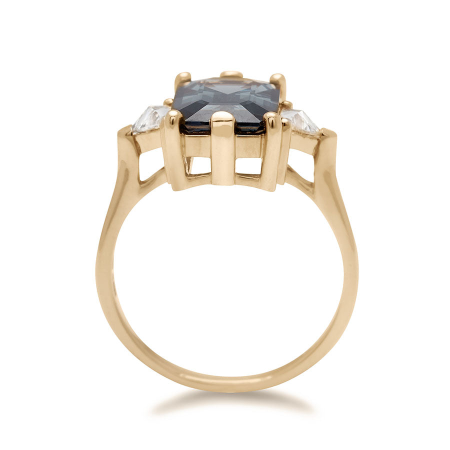 A side view of a three stone engagement ring with an emerald shape grey spinel center stone with two white diamond trillion side stones set in yellow gold.