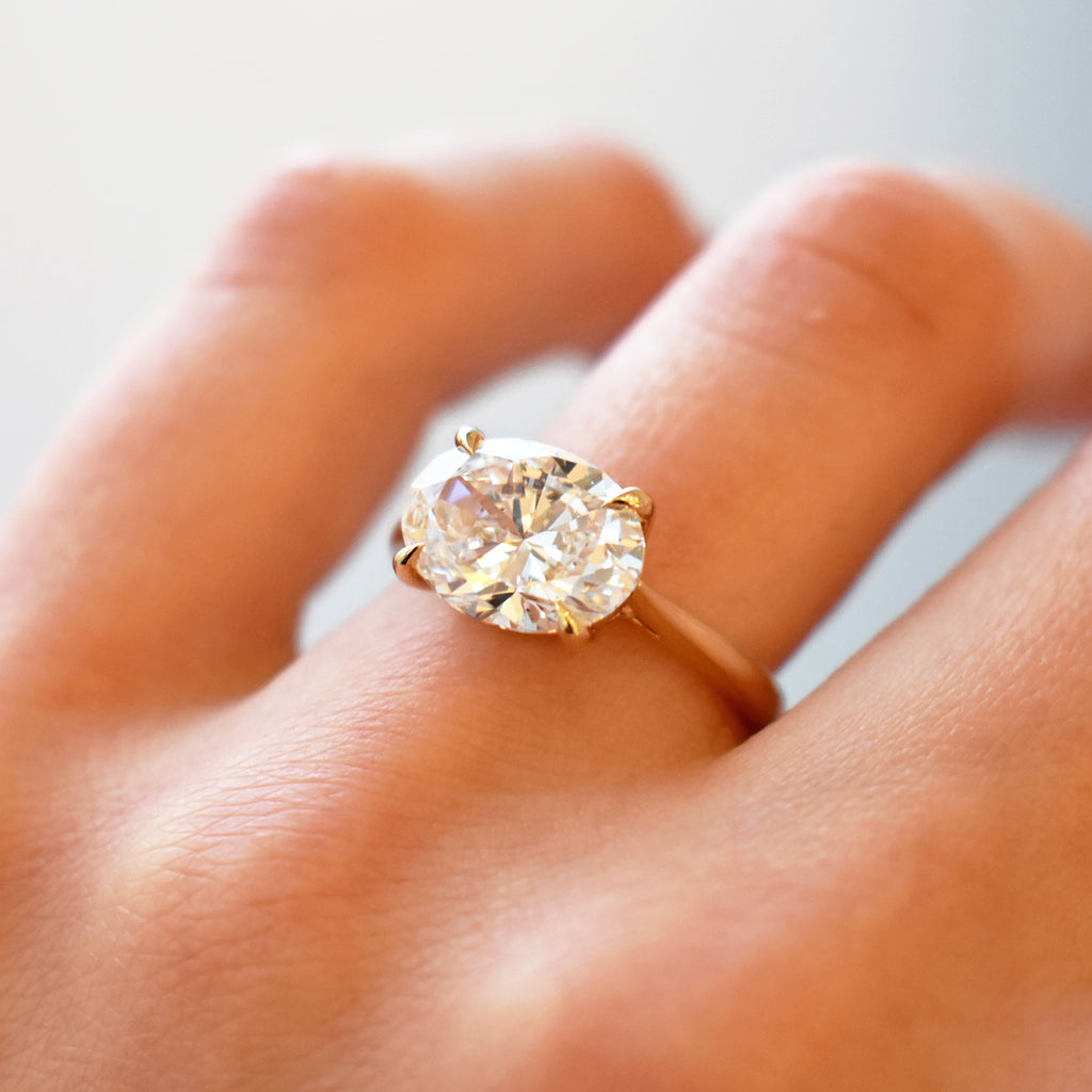 An east/west solitaire ring with an oval shape white diamond stone set in yellow gold on a light-complextion hand.