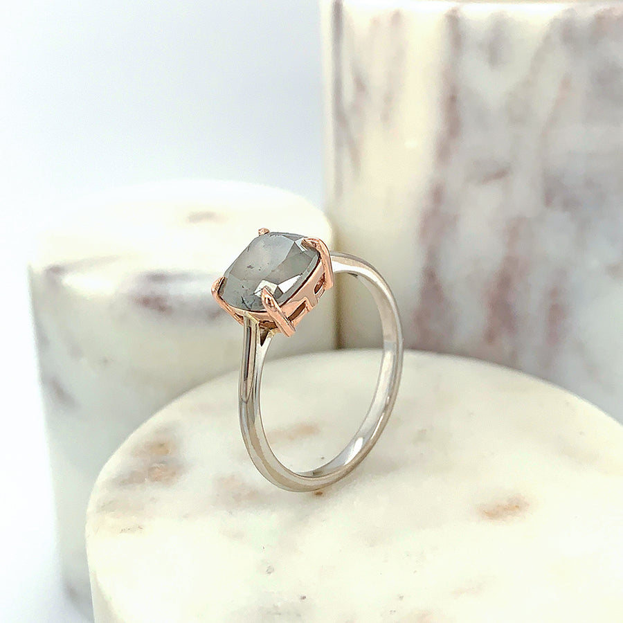 An east/west solitaire ring with an oval shape grey diamond stone set in a rose gold basket and a white gold band on a marble stone.