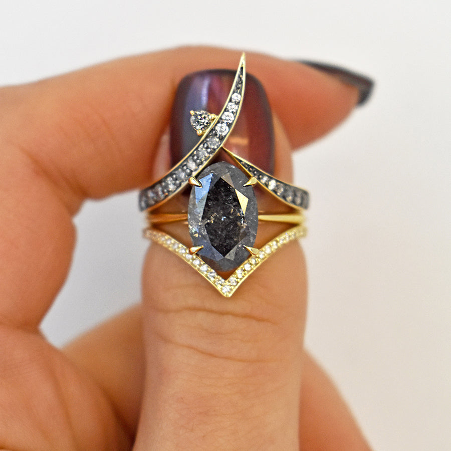 A solitaire ring oval grey diamond set in yellow gold set above a diamond dusted chrysalis shaped band and a wrap band on a light-complextion thumb.