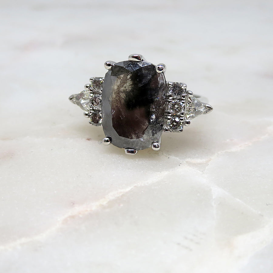 A emerald shape grey diamond center stone ring with three round white diamonds on either and two white diamond trillions on the outer edges of each side set in white gold on a grey background.