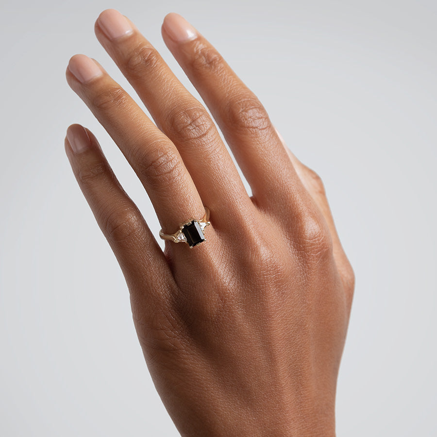 A three stone engagement ring with an emerald cut and emerald shape 8x6mm black diamond center stone with two white diamond trillion side stones set in yellow gold on a medium-complexion hand., White Gold, Rose Gold, Yellow Gold