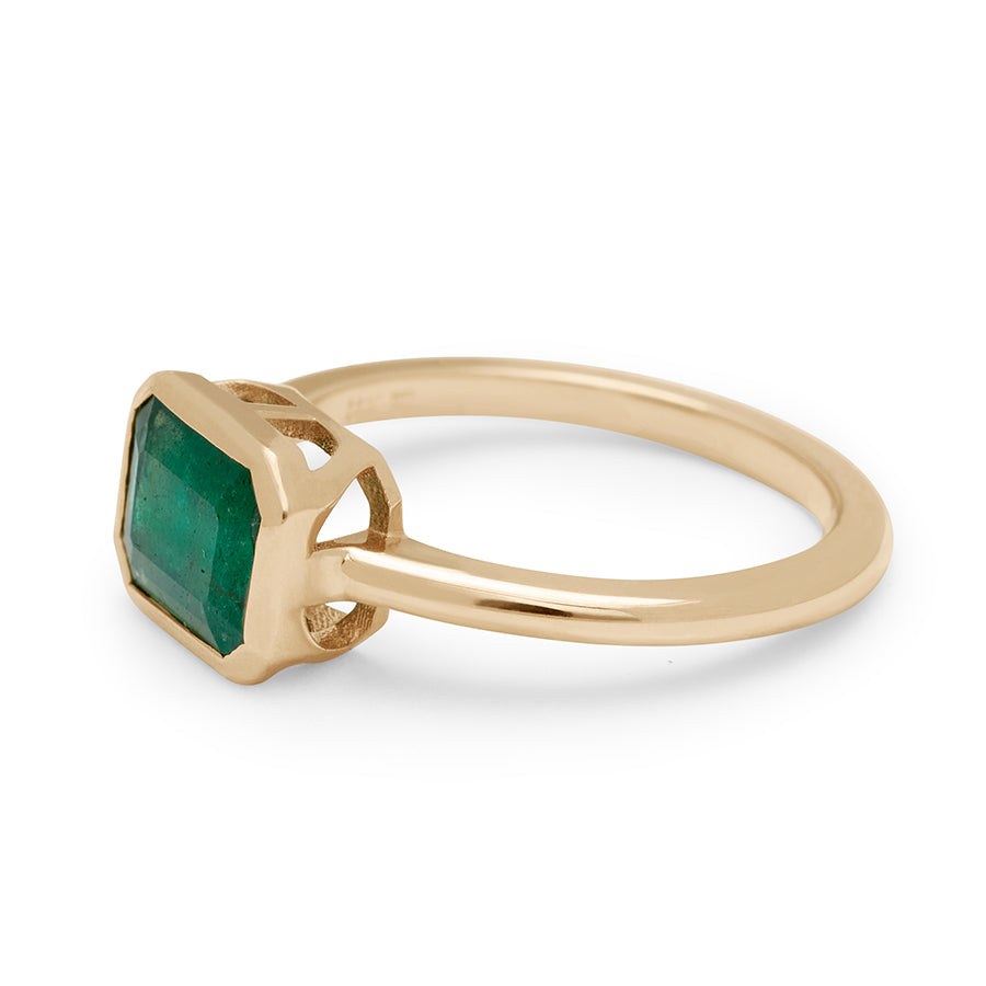 Angled view of an east/west emerald shape bezel set green emerald center set in yellow gold.