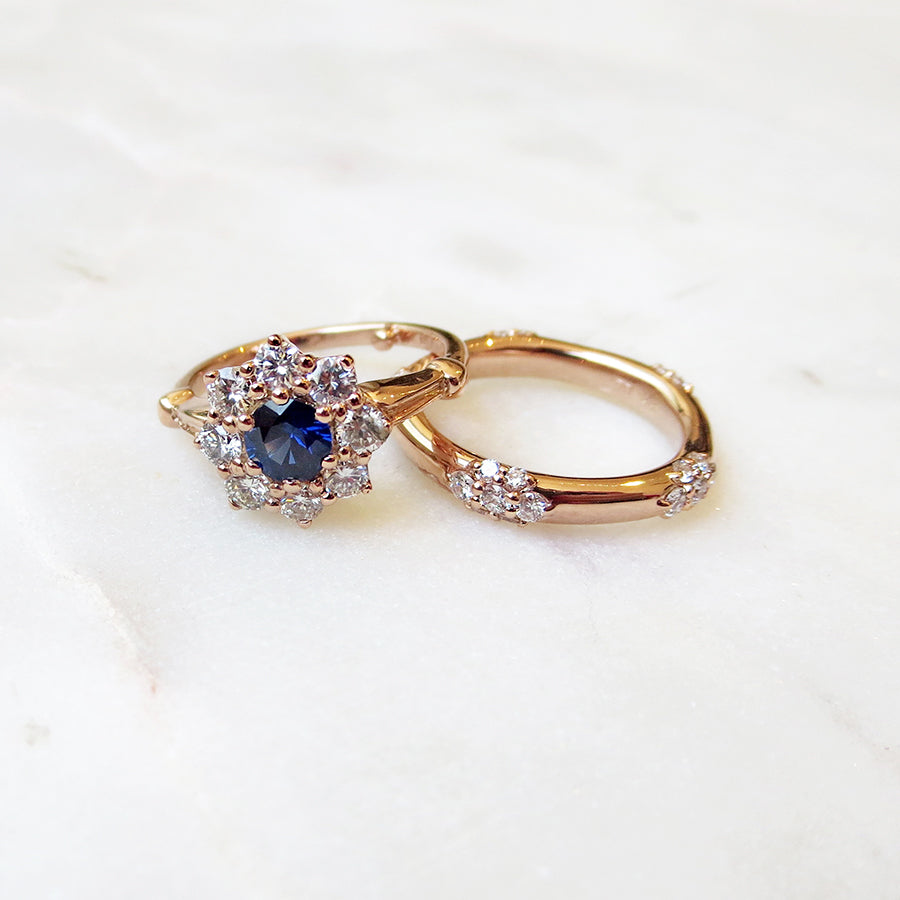A cluster engagement ring with a rose cut blue sapphire center stone and round white diamond stones surrounding it set in yellow gold with a five cluster white diamond yellow gold band on a grey background.