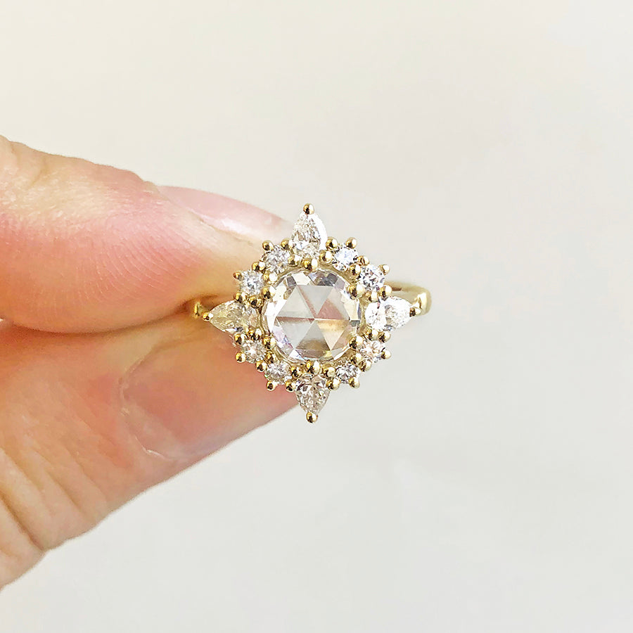A light complexion thumb and pointer finger holding a cluster engagement ring with a rose cut white diamond center stone and round white diamond stones surrounding it set in yellow gold on a grey background.