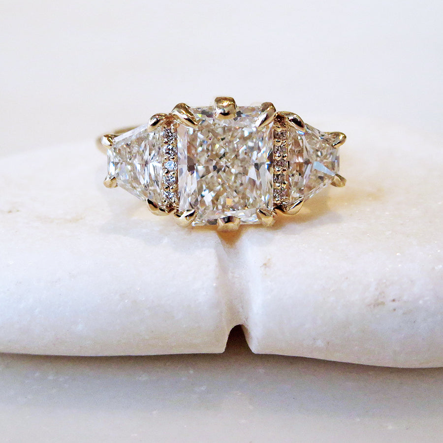 A emerald shape white diamond center stone ring with three round white diamonds on either and two white diamond trillions on the outer edges of each side set in yellow gold on a grey background.