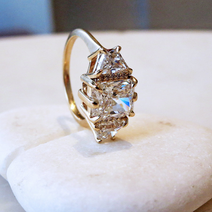 A emerald shape white diamond center stone ring with three round white diamonds on either and two white diamond trillions on the outer edges of each side set in yellow gold on a grey background.