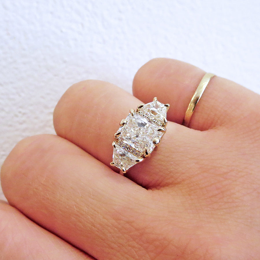 A emerald shape white diamond center stone ring with three round white diamonds on either and two white diamond trillions on the outer edges of each side set in yellow gold on a medium-complextion hand.