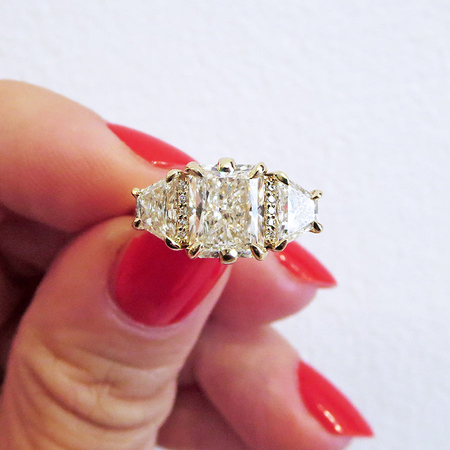 A emerald shape white diamond center stone ring with three round white diamonds on either and two white diamond trillions on the outer edges of each side set in yellow gold on a medium-complextion hand with red neails