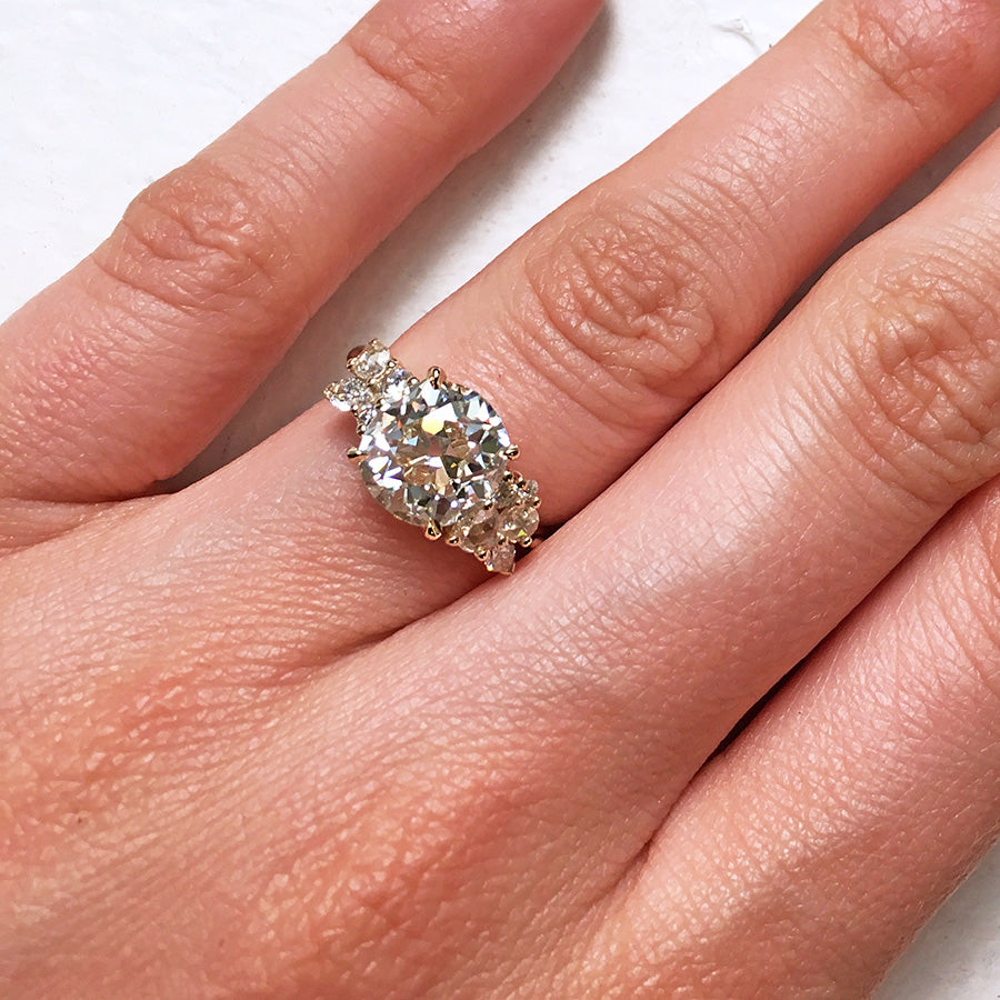 A ring with a round white diamond center stone with two clusters of white diamonds on either side set in yellow gold on a light-complexion hand.