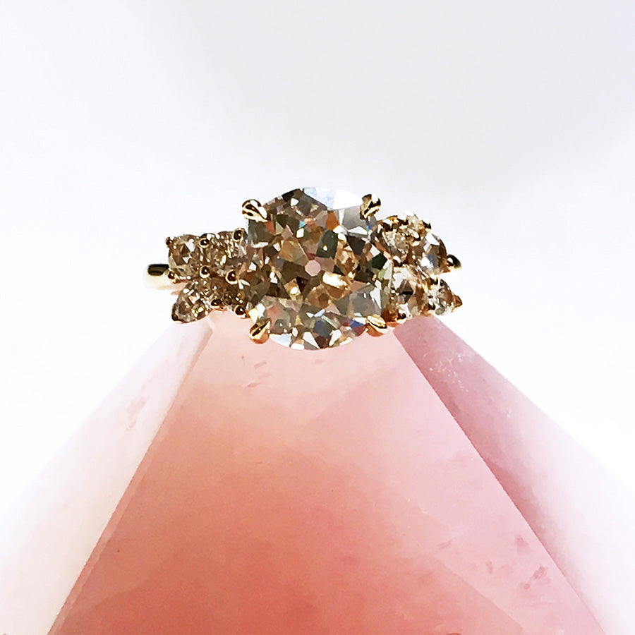 A ring with a round white diamond center stone with two clusters of white diamonds on either side set in yellow gold on a pink stone.
