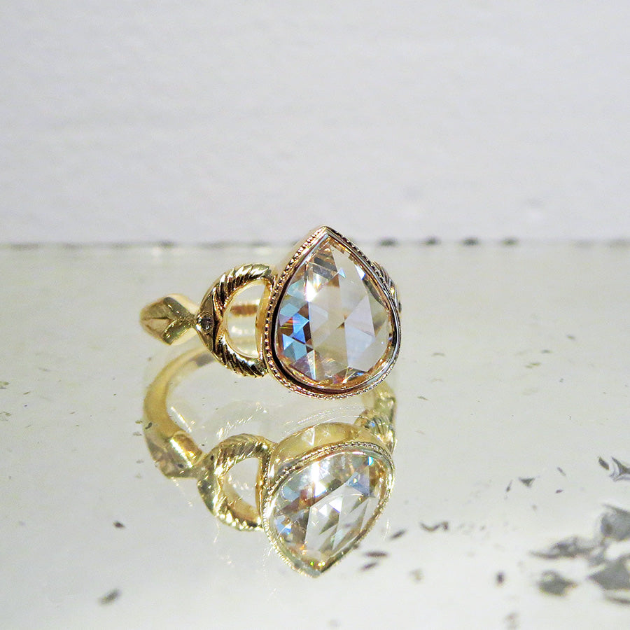 Angled view of a pear rose cut white diamond with engraved gold accents on either side set in yellow gold on a mirror.