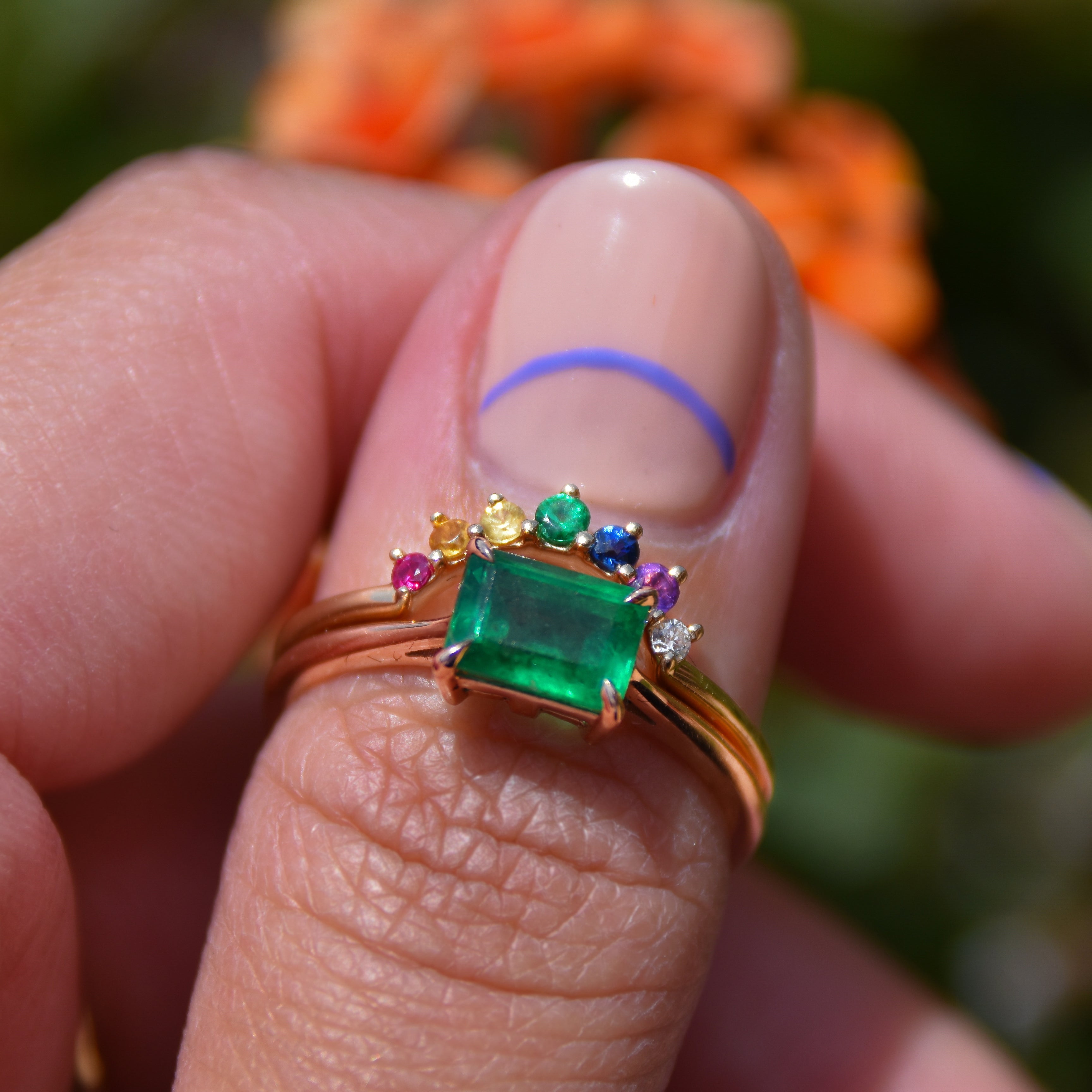 A curved 7 rainbow colored gemstones band set in yellow gold stacked above a east/west emerald shape green emerald ring on a light-compelxion thumb.