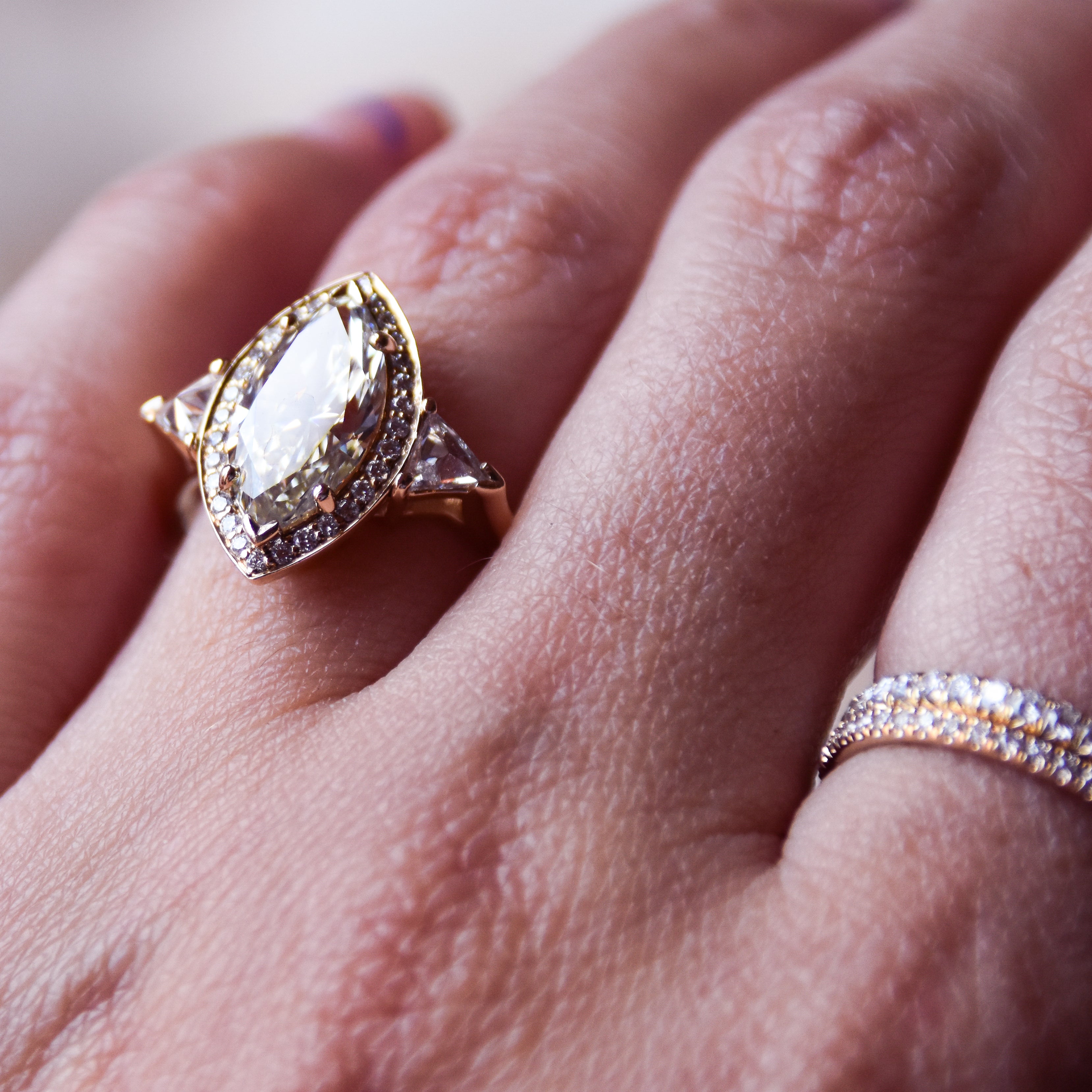 A three stone engagement ring with a marquise shape champagne diamond center stone surrounded by a white diamond pave halo with two white diamond trillion side stones set in yellow gold on a ring finger with a diamond dusted straight bands on the pointer finger.