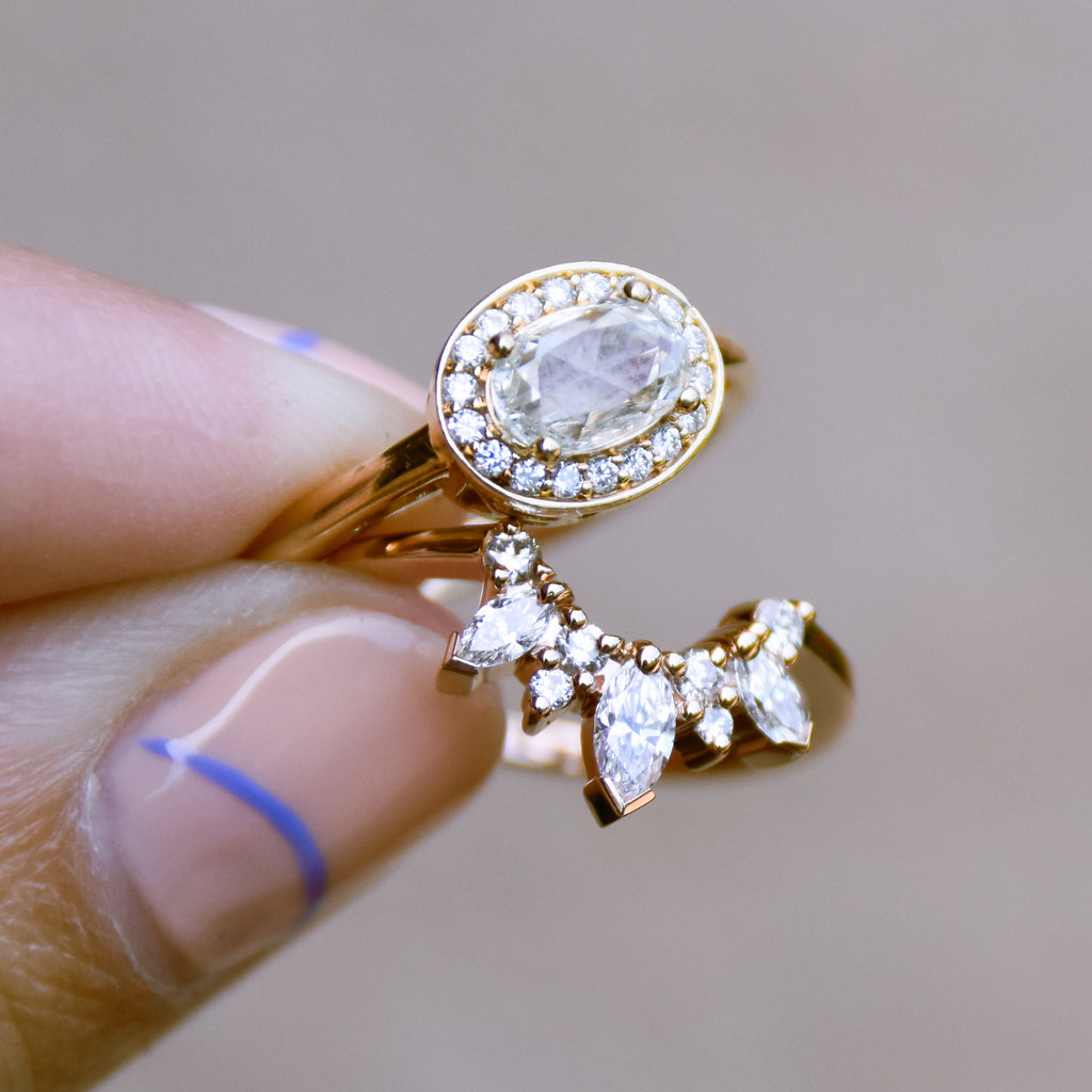 A hand holding a band with three marquise shaped white diamonds with round white diamond set in yellow gold and an oval halo engagement ring.