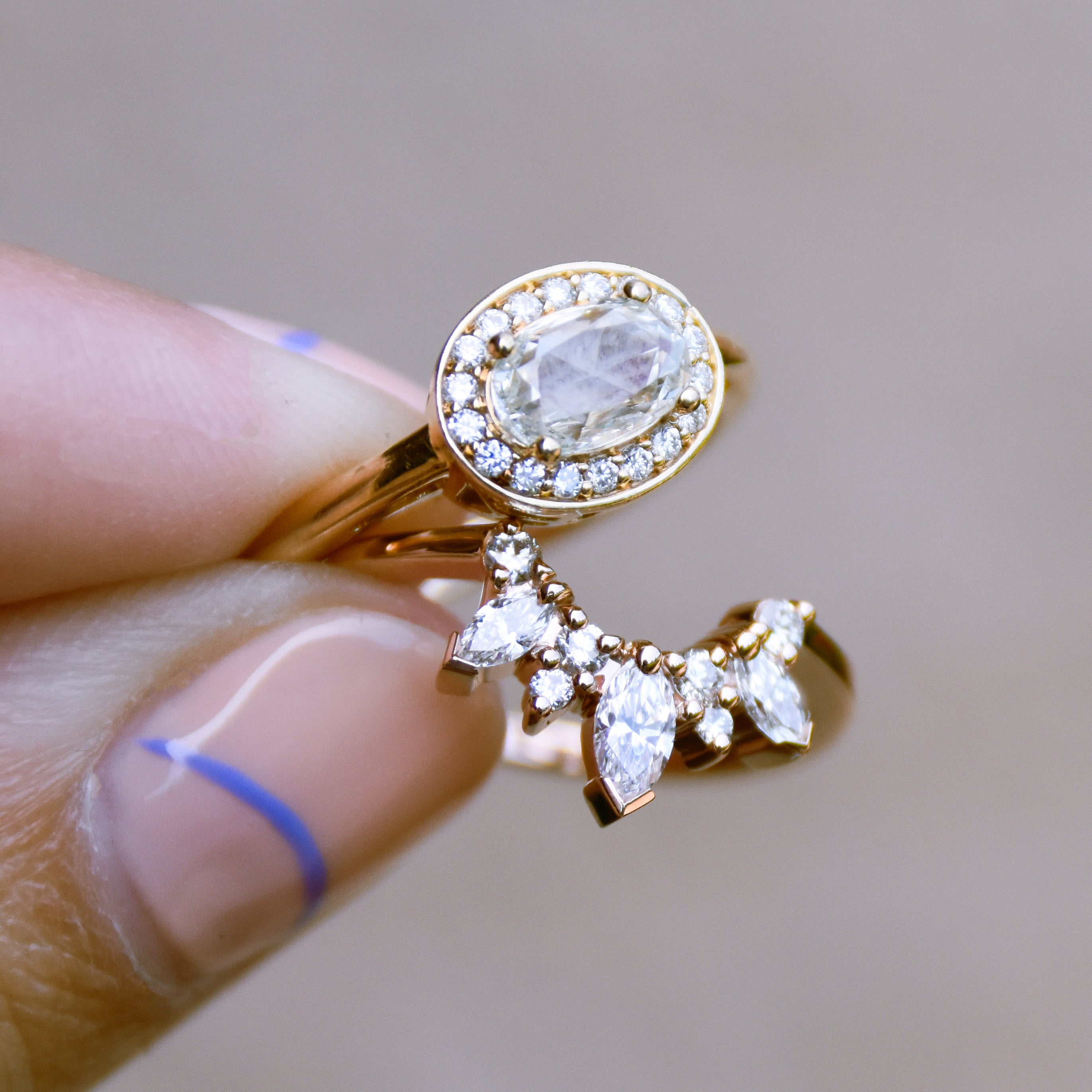 A hand holding a band with three marquise shaped white diamonds with round white diamond set in yellow gold and an oval halo engagement ring.