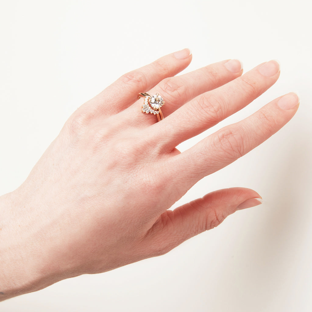 A solitaire round white diamond ring with double claw prongs set in yellow gold above a triple baguette white diamond band in yellow gold on a light-complexion hand.